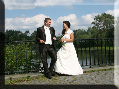 Para młoda w strojach ślubnych, stojąca przy barierce z widokiem na jezioro, trzymająca czerwoną różę, w tle drzewa i błękitne niebo z chmurami.