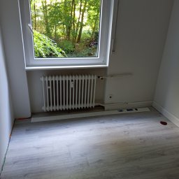 Daniel Starski Usługi Ogólnoremontowe i Ogrody - Pokój po malowaniu: białe ściany, grzejnik pod oknem z widokiem na zieleń, szare panele podłogowe. Widoczne listwy przypodłogowe i rury grzewcze.