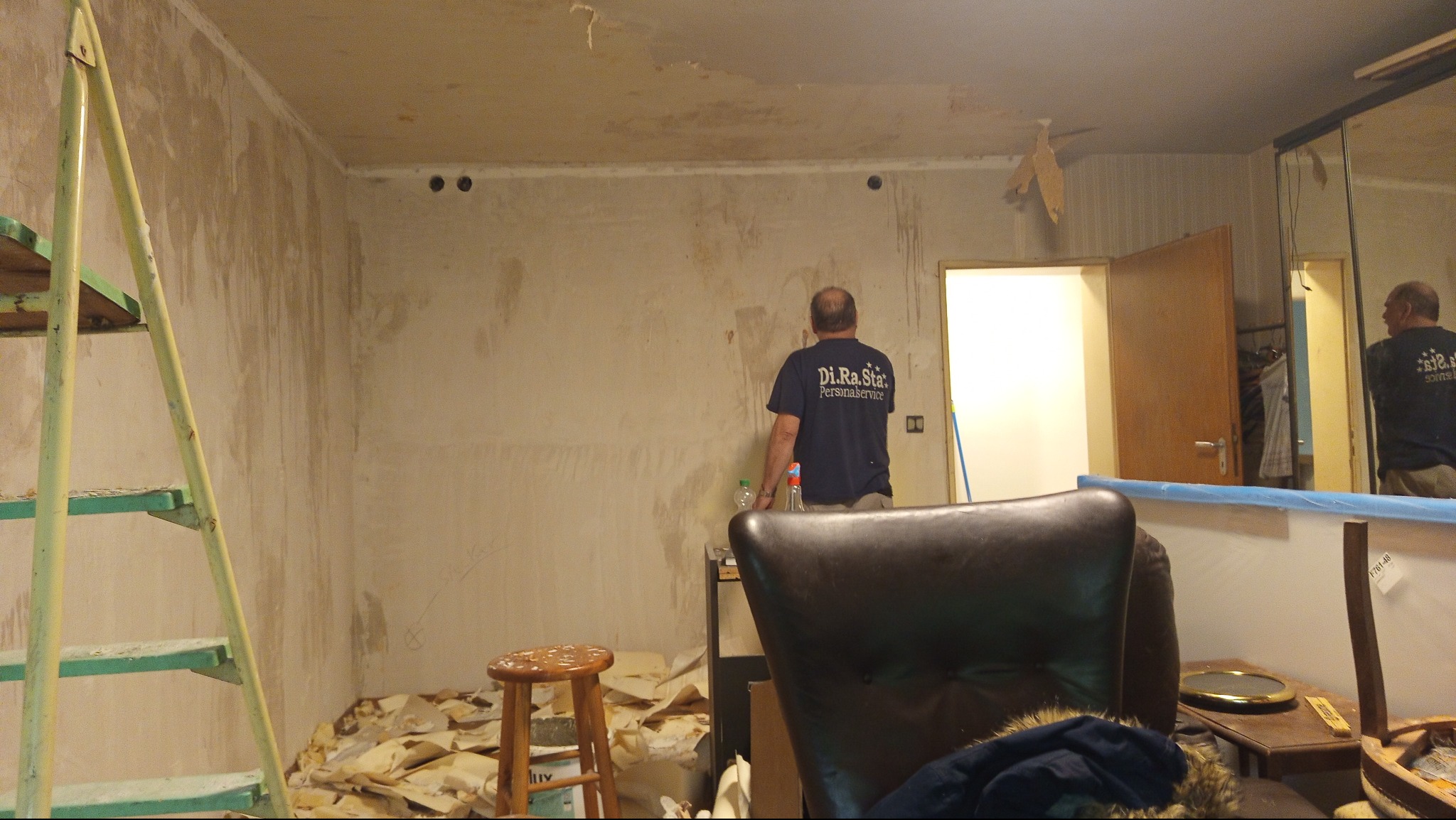 Przygotowanie ścian do malowania: usunięte stare tapety, widoczny pracownik w pomieszczeniu z drabiną i stołkiem. Ściany wymagają dalszej obróbki.