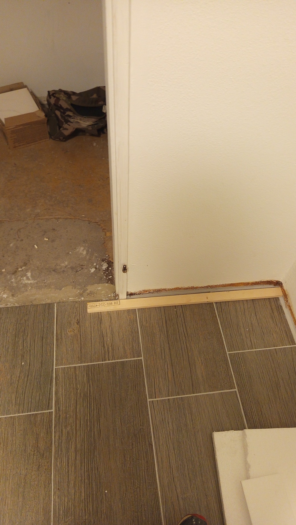 Fragment wnętrza w trakcie remontu: brązowe płytki podłogowe imitujące drewno, biała ściana i próg drzwiowy z widocznymi śladami po starej farbie.