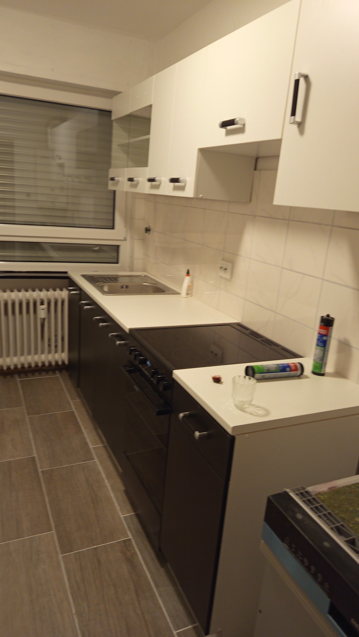 Nowoczesna kuchnia po remoncie: białe szafki wiszące, ciemne szafki dolne, jasny blat, okno z żaluzjami. Widoczne akcesoria kuchenne i płytki na ścianie.