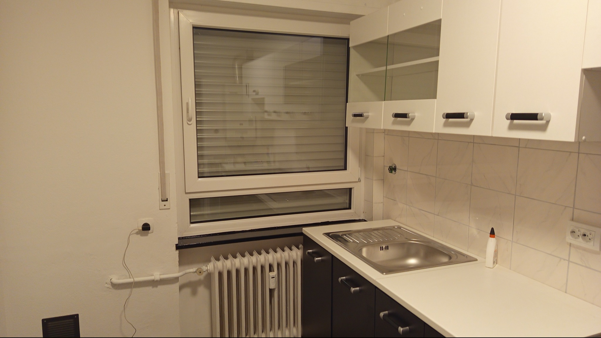 Jasna kuchnia po remoncie: białe ściany, okno z żaluzjami, ciemne szafki pod blatem i białe wiszące szafki. Widoczny grzejnik i płytki na ścianie.