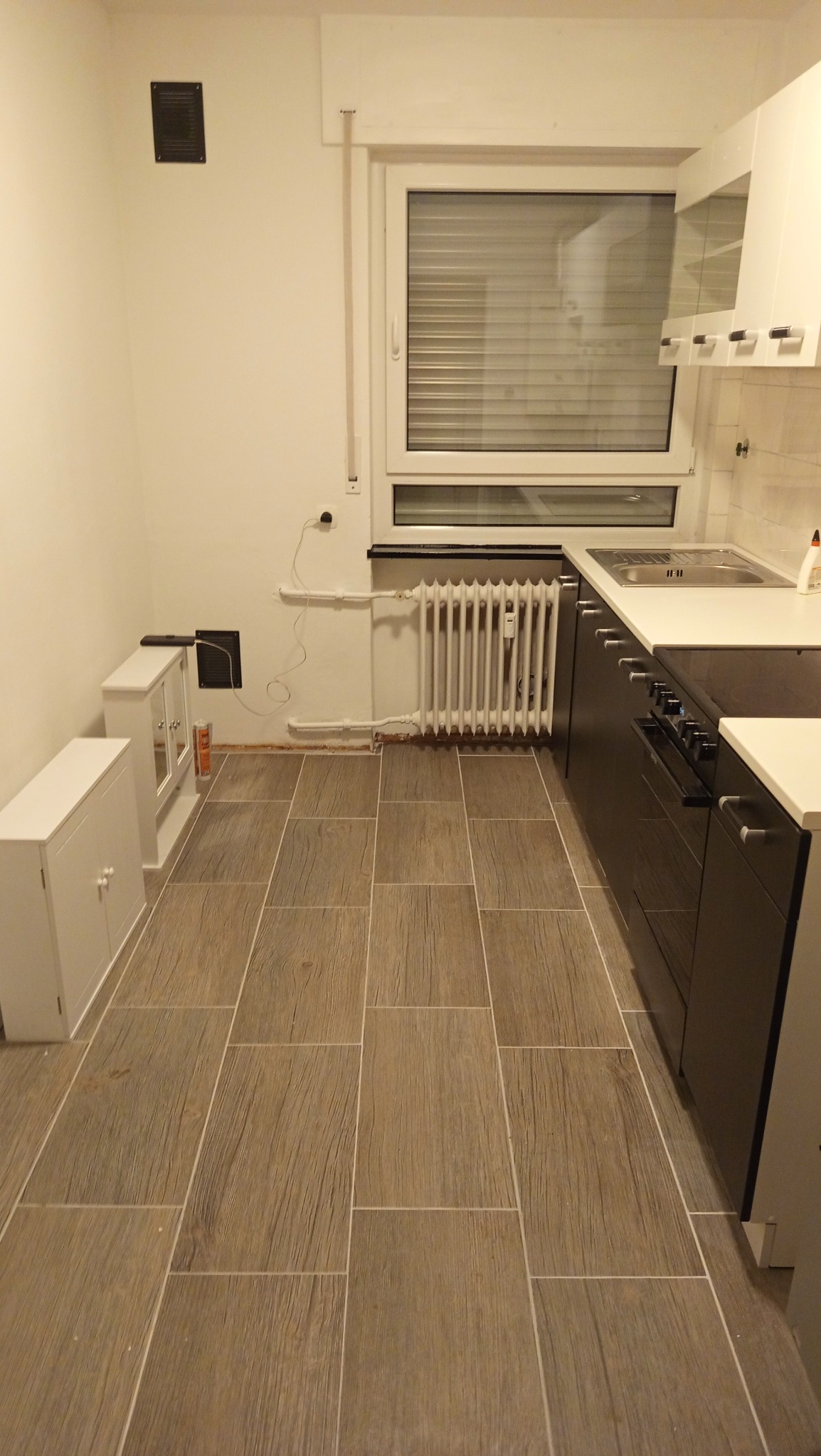 Jasna kuchnia po remoncie: białe ściany, brązowe płytki imitujące drewno, ciemne szafki kuchenne. Widoczny grzejnik i okno z żaluzjami.
