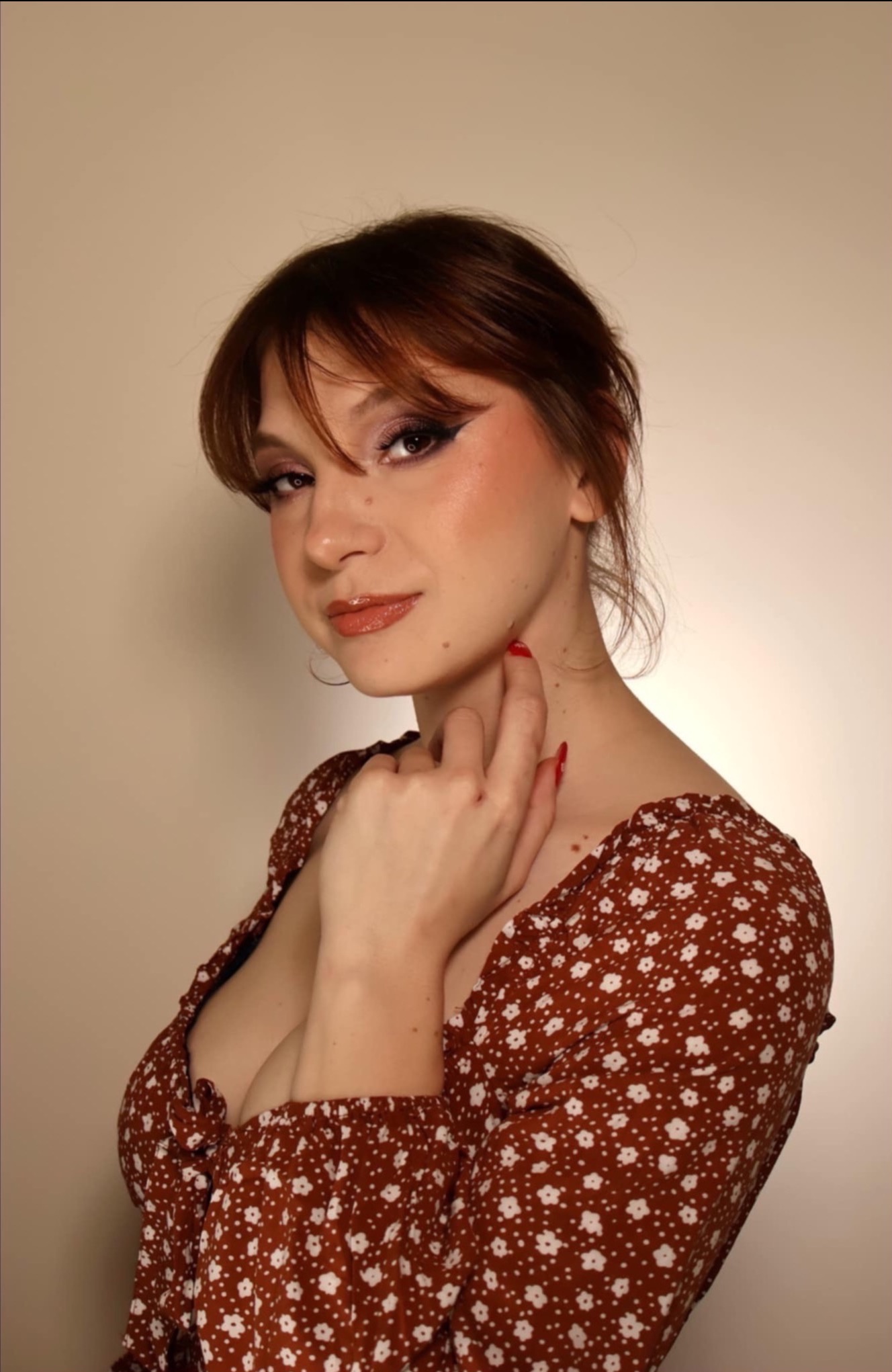 Portret kobiety z delikatnym makijażem oczu i ust w odcieniach brązu, z czerwoną stylizacją paznokci, pozującej w kwiecistej sukience na jasnym tle.