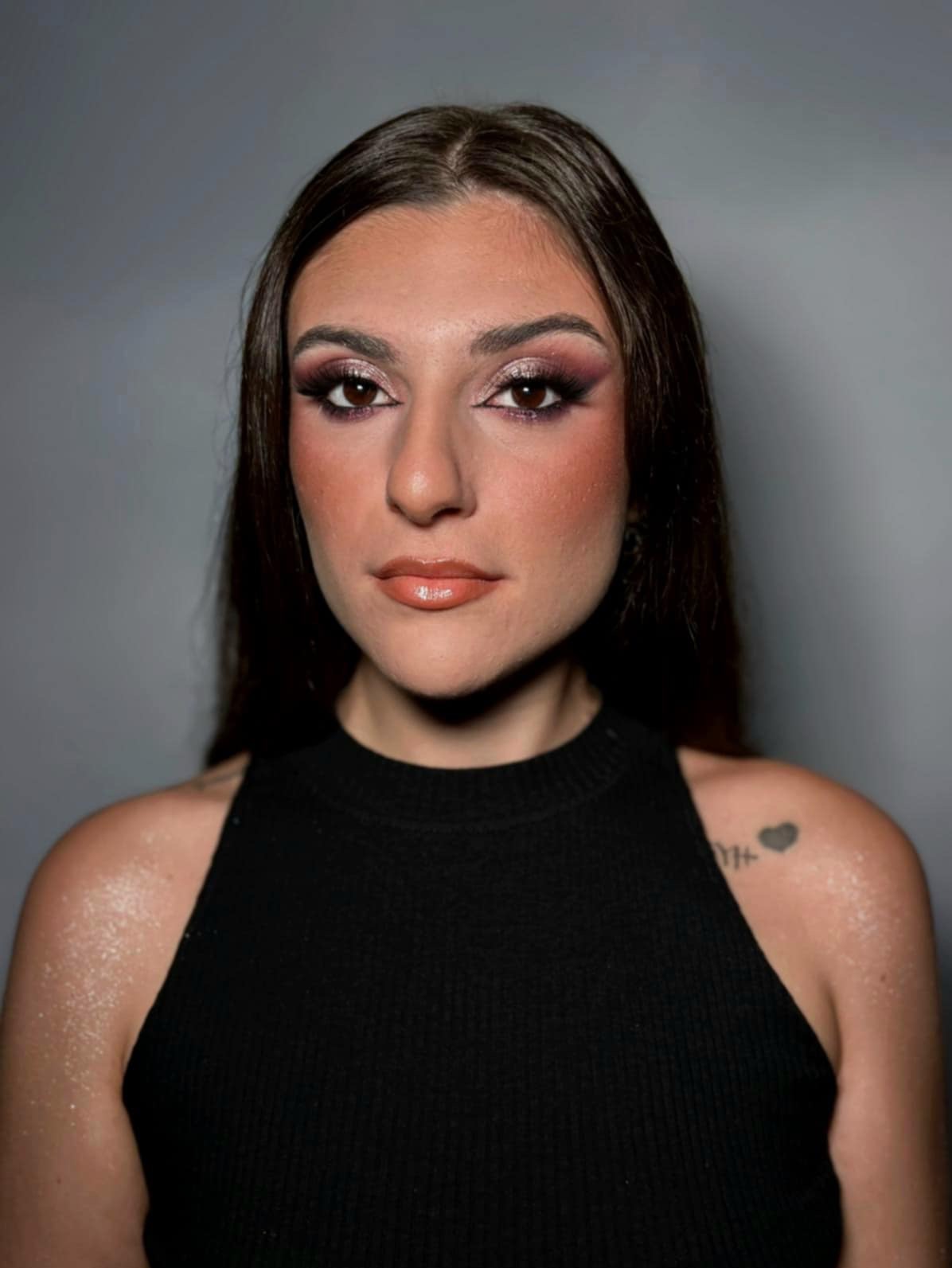 Portret kobiety z intensywnym makijażem oczu w odcieniach brązu i fioletu, błyszczące usta, ciemne włosy i tatuaż serca na ramieniu na szarym tle.