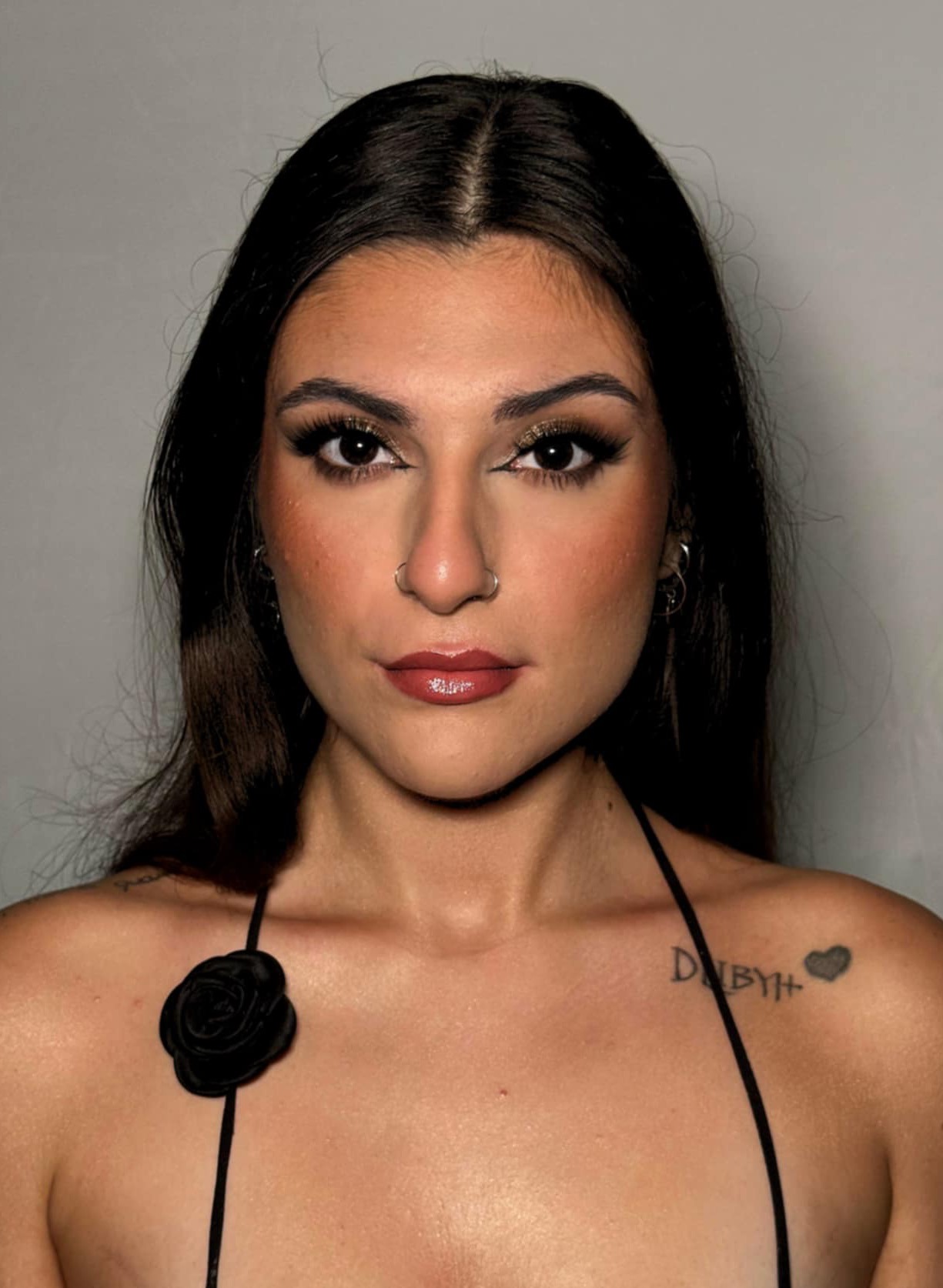 Portret kobiety z ciemnymi włosami, w wyrazistym makijażu oczu z brokatem i błyszczącymi ustami. Widoczny tatuaż na ramieniu i kolczyk w nosie.