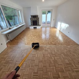 Tad-pol - Parkiet po renowacji, pokryty lakierem. Widoczny wałek do lakierowania, fragment dłoni. W tle kominek i okno. Jasne wnętrze po remoncie.