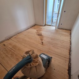 Tad-pol - Cyklinowanie drewnianej podłogi w pokoju, widoczna maszyna do cyklinowania i częściowo wyszlifowana powierzchnia. Okno zabezpieczone folią malarską.