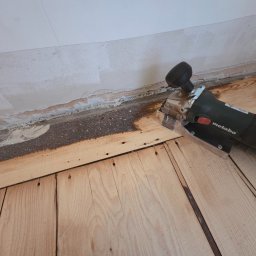 Tad-pol - Elektryczna frezarka Metabo przy krawędzi drewnianej podłogi, przygotowywanej do cyklinowania. Widoczne ślady po starym wykończeniu przy ścianie.