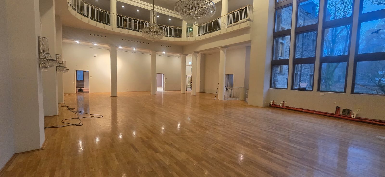 Duża sala z parkietem po renowacji, kolumny, żyrandole i okna z widokiem na zewnątrz. Widoczne ślady po pracach wykończeniowych.