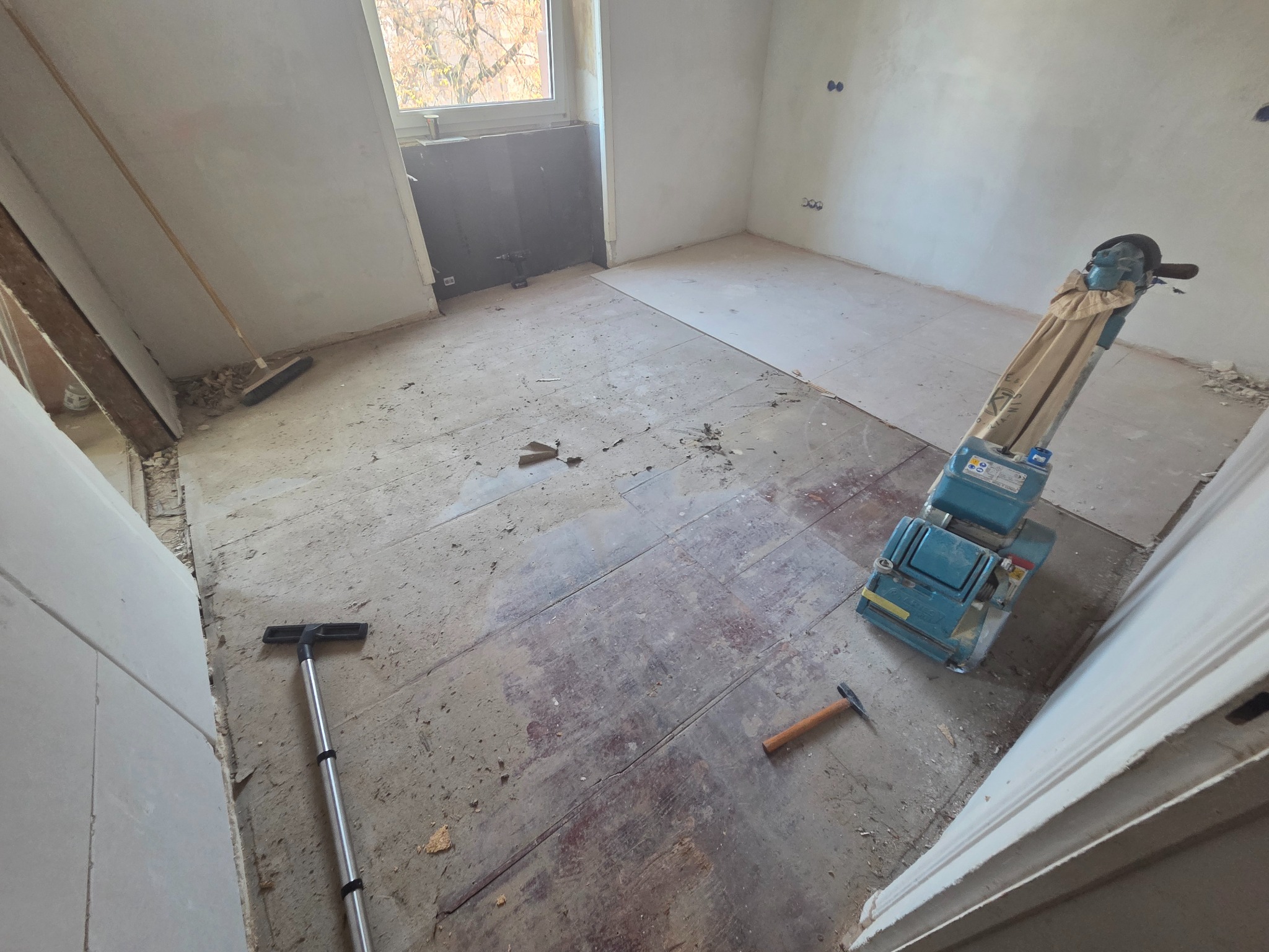 Pokój w trakcie renowacji podłogi: cykliniarka, odkurzacz i młotek na starym parkiecie. Widoczne ślady po cyklinowaniu, okno i fragment ściany z instalacją elektryczną.
