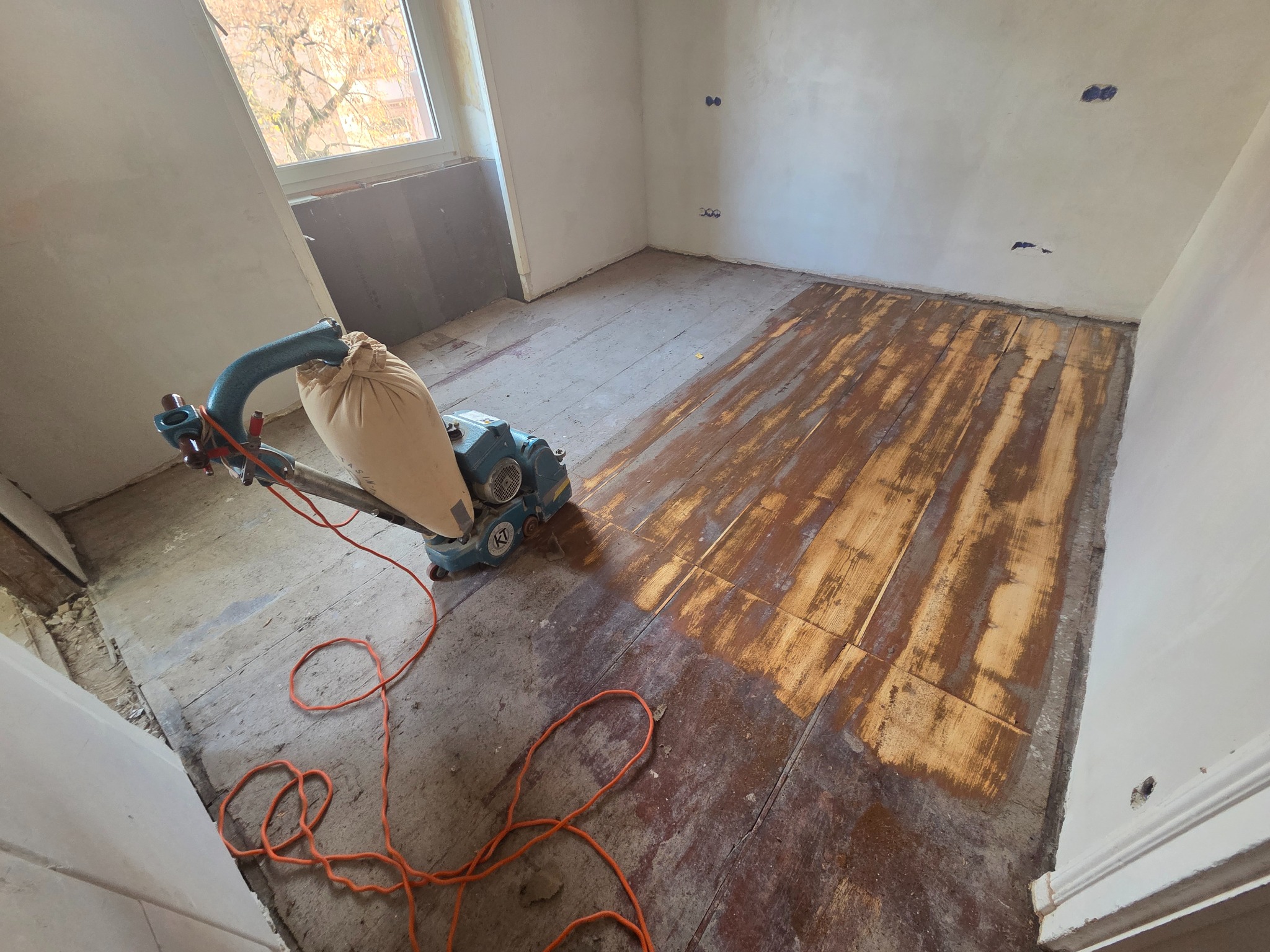 Cyklinowanie parkietu w trakcie realizacji. Widoczna maszyna do cyklinowania i fragment podłogi po obróbce, ukazujący różnicę w wyglądzie drewna. Pomieszczenie w trakcie remontu.