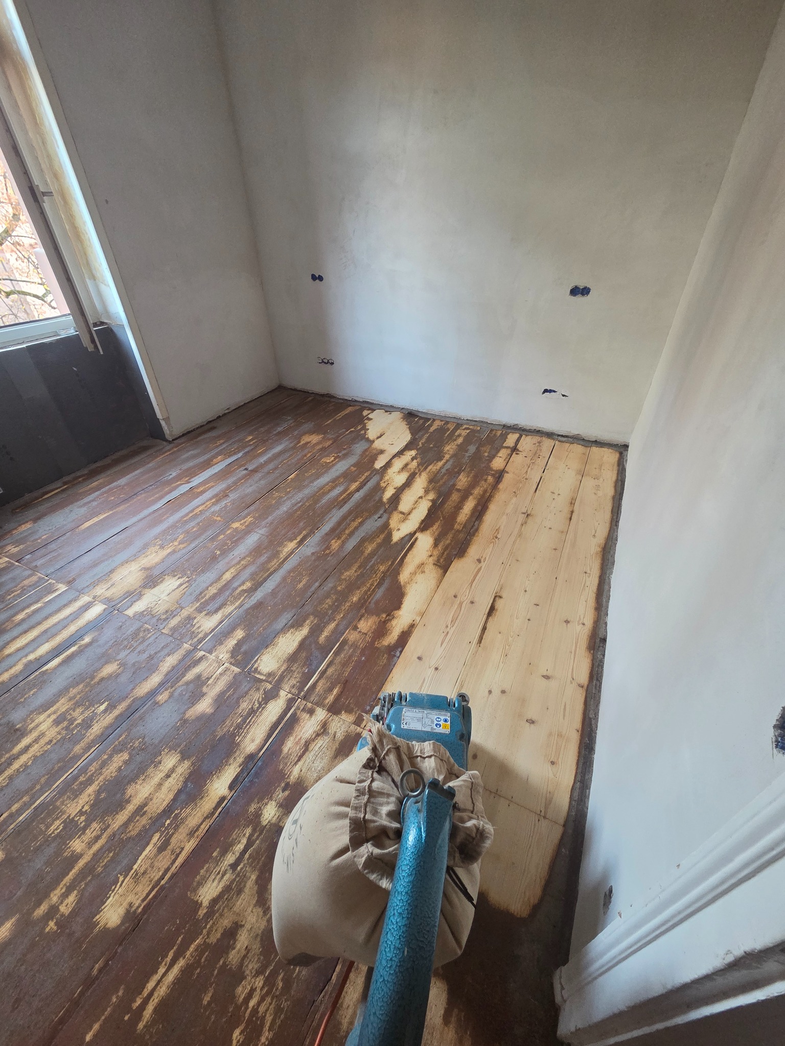 Cyklinowanie drewnianej podłogi w trakcie renowacji. Widoczna maszyna do cyklinowania i fragmenty podłogi po obróbce, ukazujące jaśniejsze drewno.