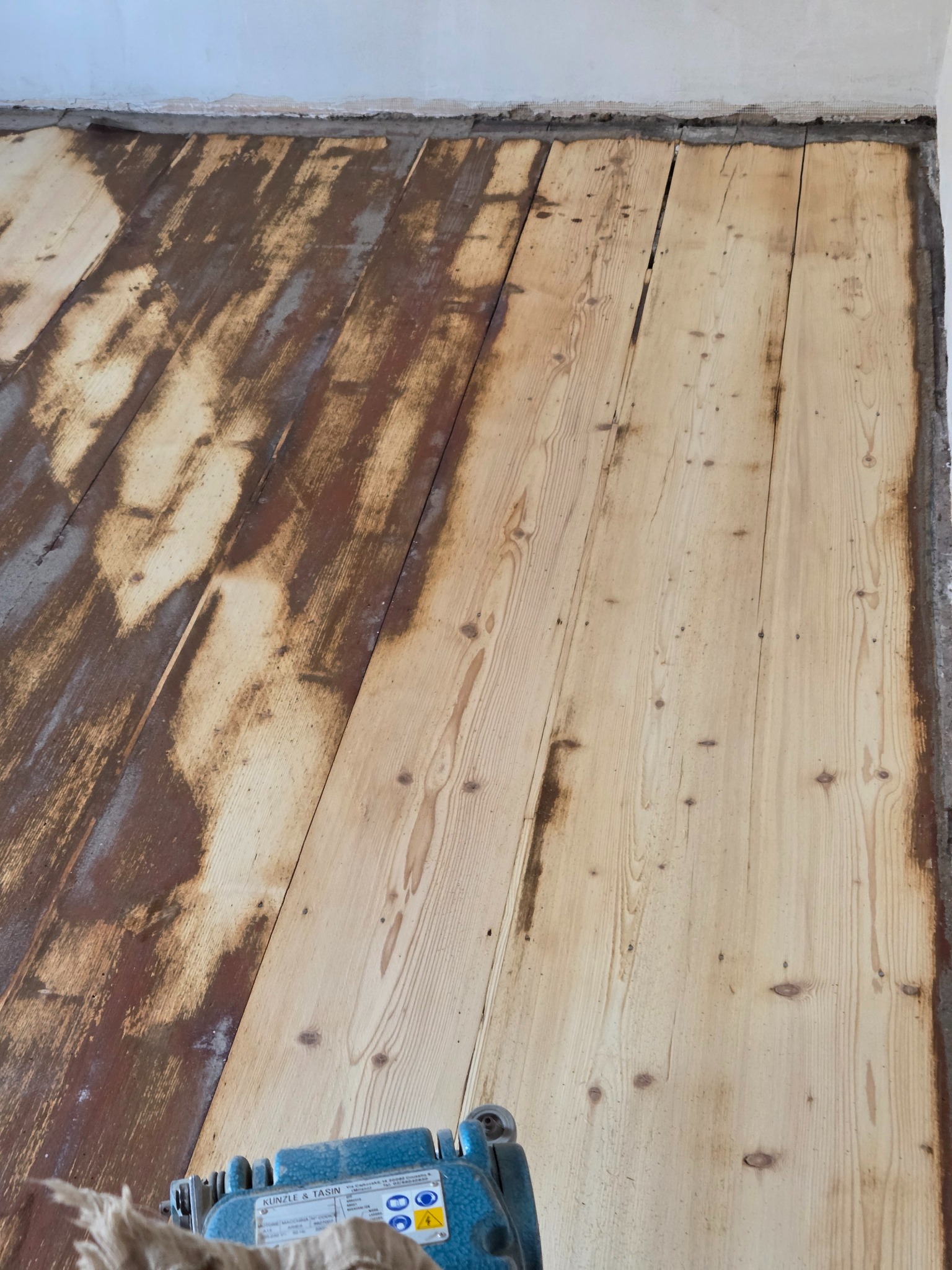 Częściowo odnowiona drewniana podłoga z widoczną maszyną do cyklinowania. Połowa podłogi w jasnym odcieniu drewna, druga w ciemniejszym, surowym stanie.