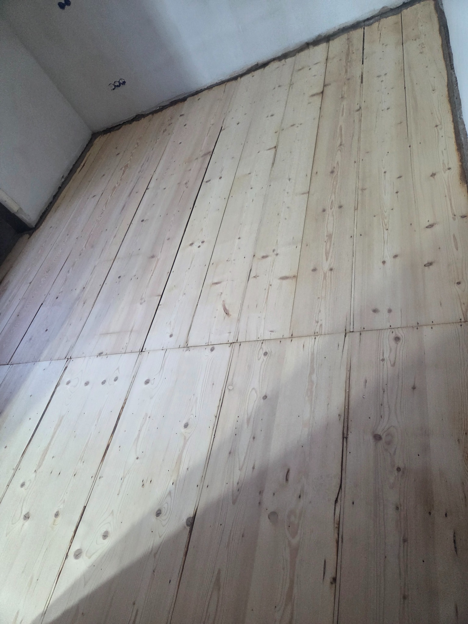 Jasne, surowe deski podłogowe ułożone wzdłużnie, widoczne ślady po gwoździach i naturalne usłojenie drewna, przygotowane do dalszej obróbki w narożnym pomieszczeniu.