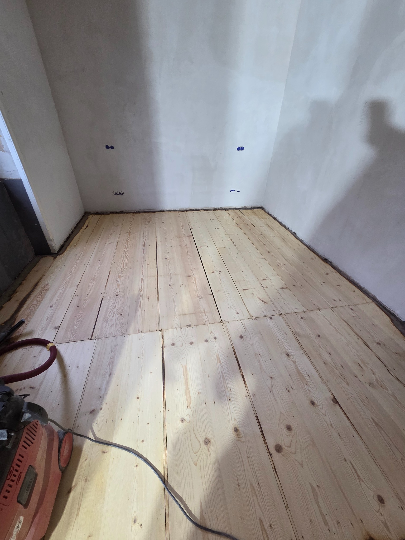 Surowe deski podłogowe w trakcie montażu w rogu pokoju, widoczne ślady po instalacji elektrycznej na ścianie i sprzęt do obróbki drewna.