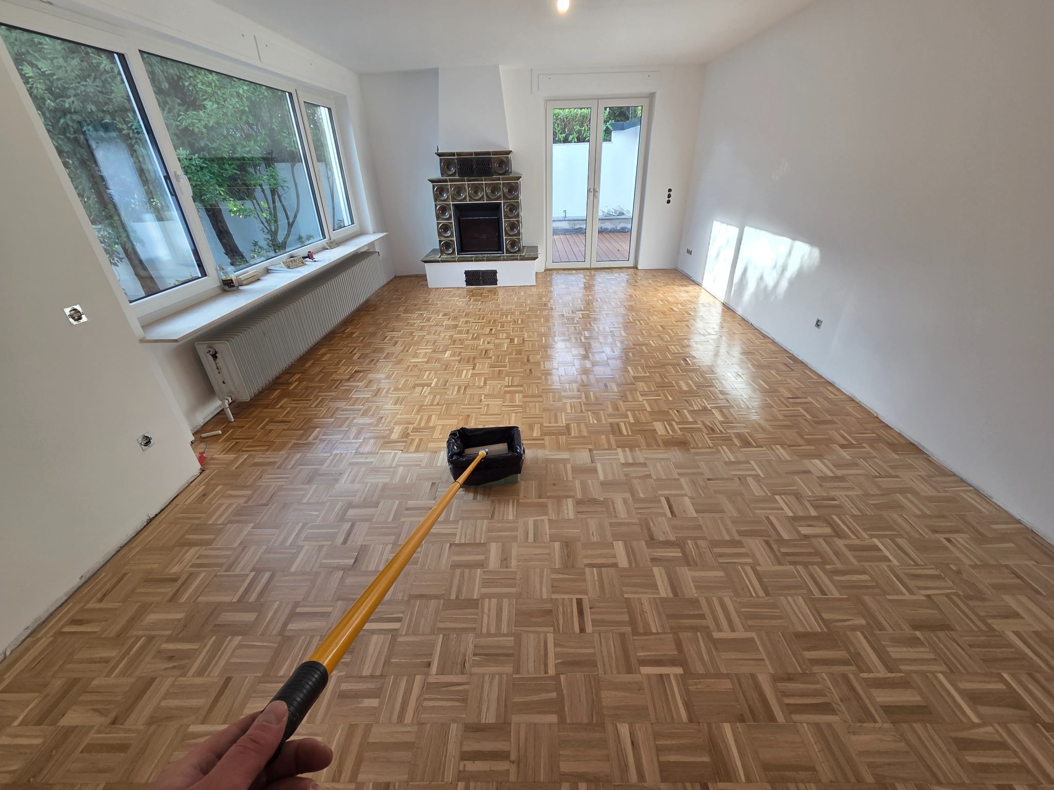 Parkiet po renowacji, pokryty lakierem. Widoczny wałek do lakierowania, fragment dłoni. W tle kominek i okno. Jasne wnętrze po remoncie.