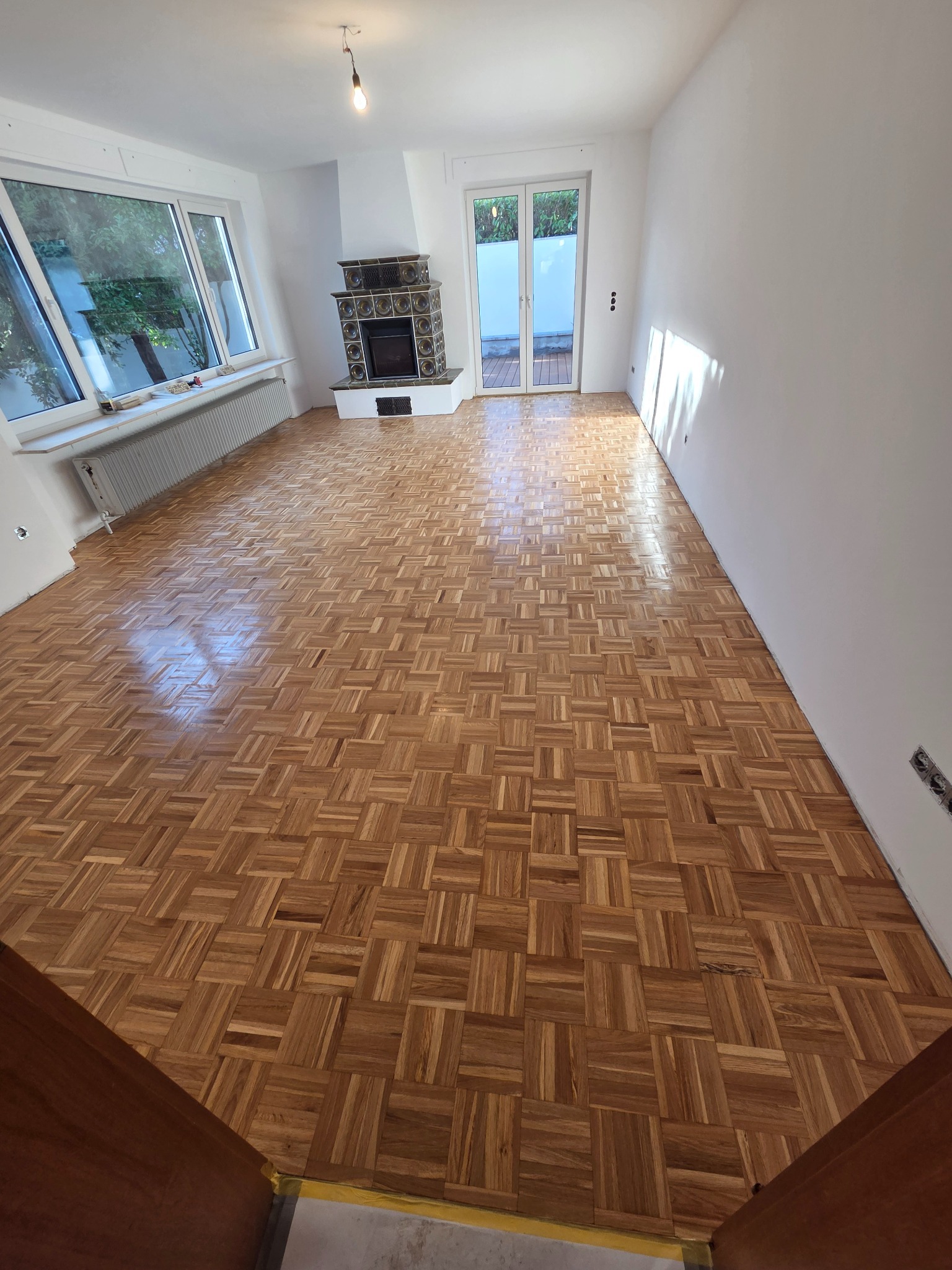 Parkiet w jodełkę po renowacji w jasnym pokoju z oknem i piecem kaflowym. Widoczne detale ułożenia i wykończenia podłogi. Perspektywa od progu.