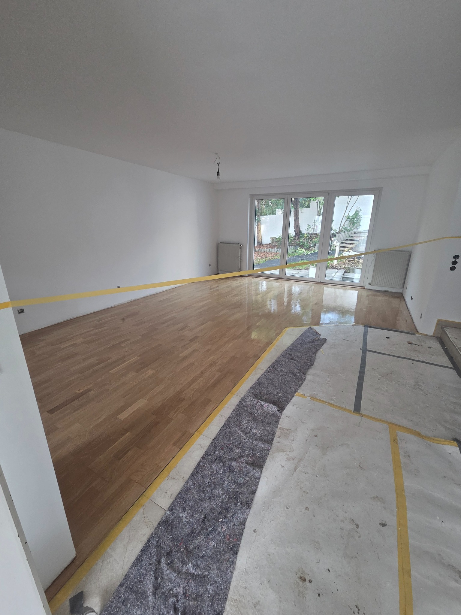 Jasny pokój z częściowo wycyklinowanym parkietem, zabezpieczonym taśmą. Fragment podłogi przygotowany do dalszych prac, przykryty szarą tkaniną. Duże okno balkonowe.