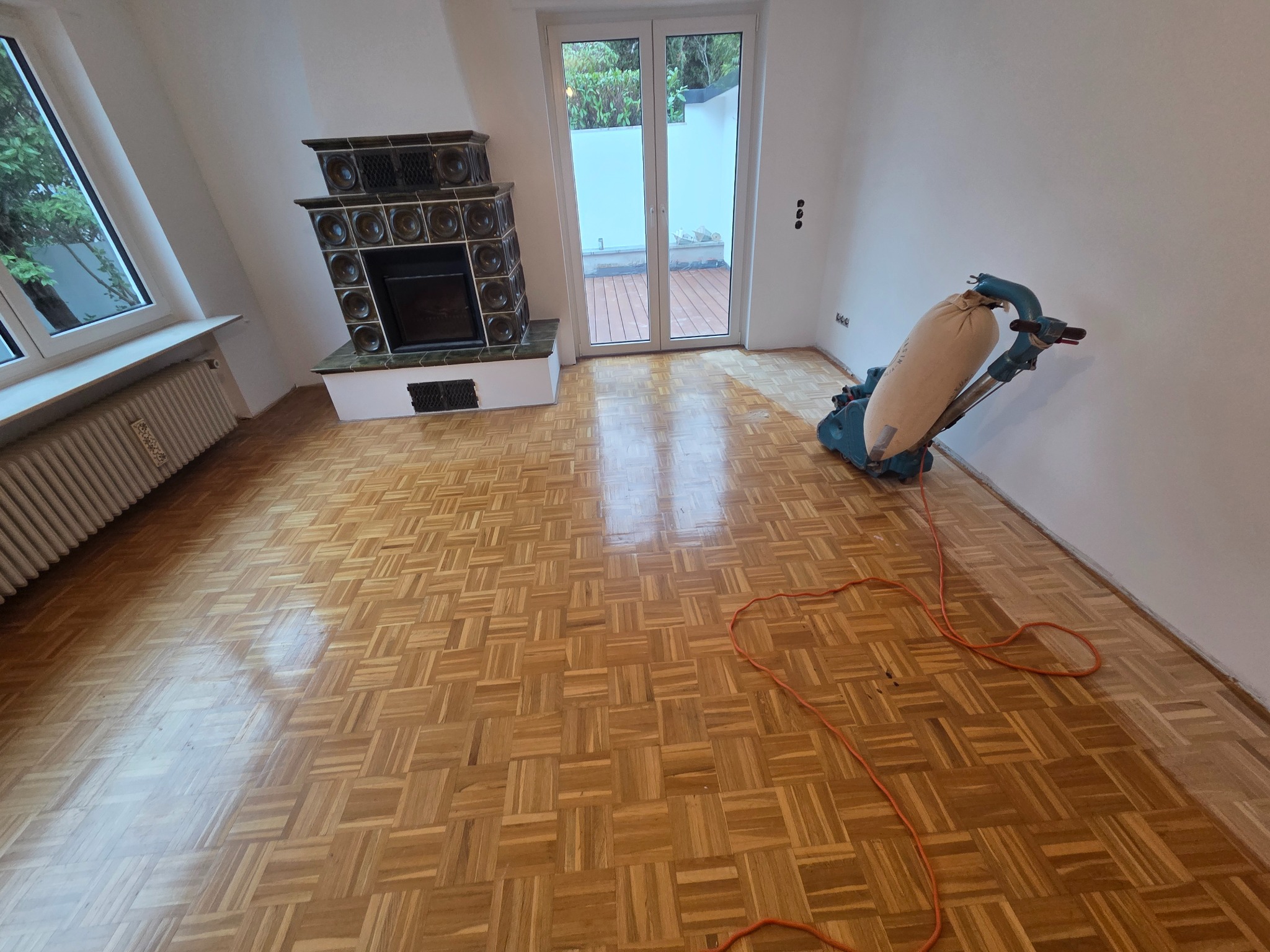 Cyklinowanie parkietu w pokoju z kominkiem i wyjściem na taras. Widoczna maszyna do cyklinowania i świeżo wycyklinowany parkiet w jodełkę.