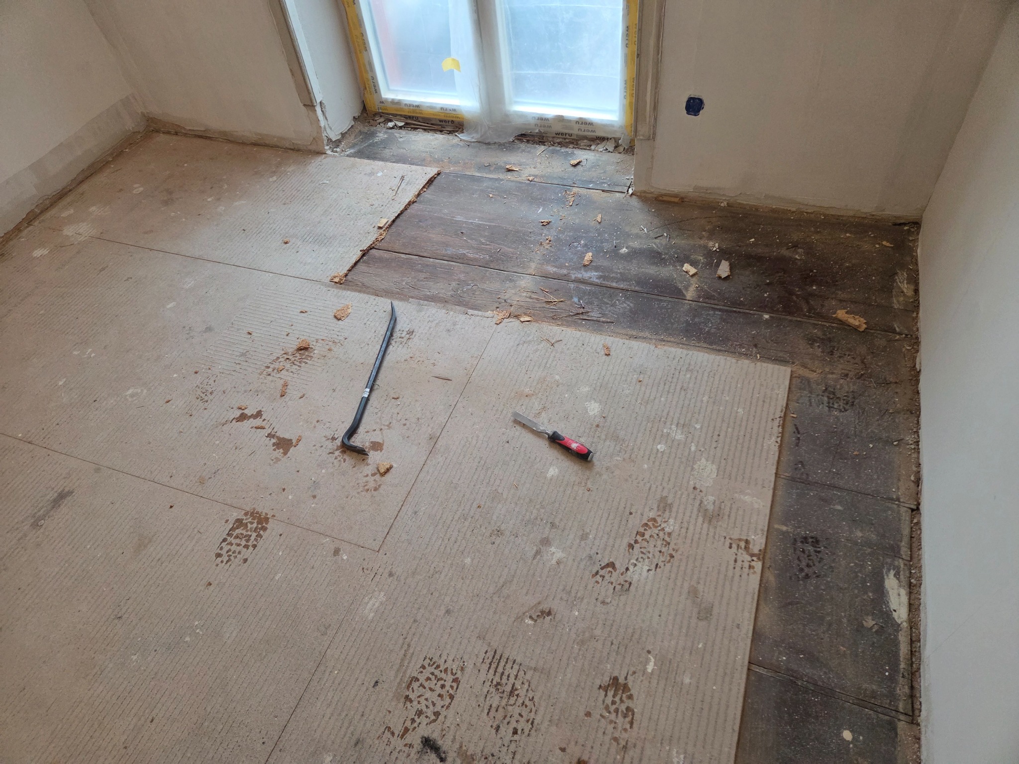 Demontaż starej podłogi w pokoju: fragmenty drewnianej podłogi, płyty podkładowe, ślady butów, łom i dłuto. Widok z góry na remontowane wnętrze.