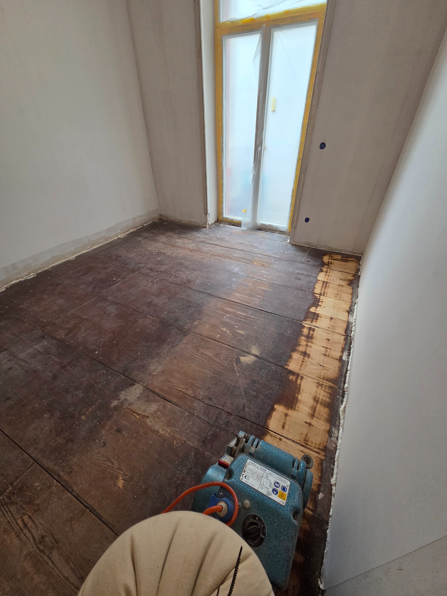 Cyklinowanie podłogi w trakcie realizacji. Część podłogi wycyklinowana, widoczna maszyna do cyklinowania. Okno zabezpieczone folią malarską.