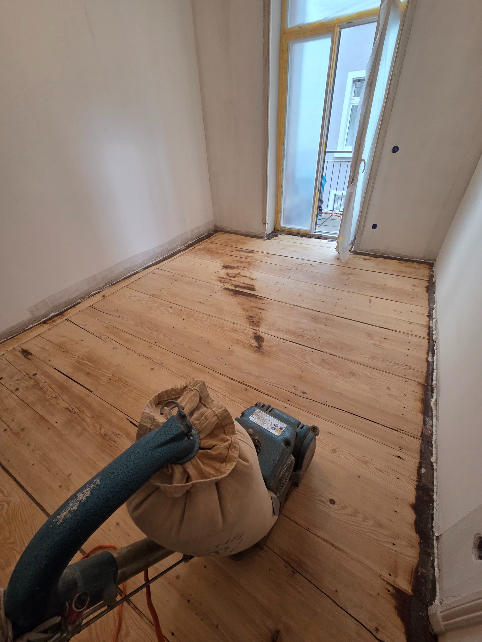 Cyklinowanie drewnianej podłogi w pokoju, widoczna maszyna do cyklinowania i częściowo wyszlifowana powierzchnia. Okno zabezpieczone folią malarską.
