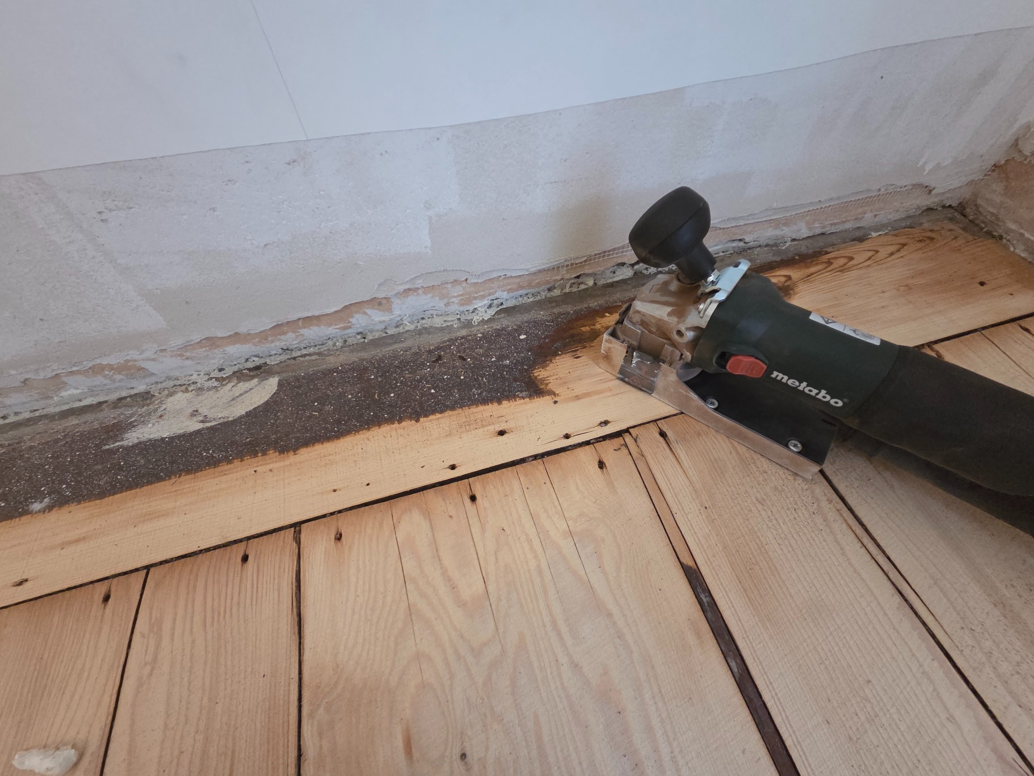Elektryczna frezarka Metabo przy krawędzi drewnianej podłogi, przygotowywanej do cyklinowania. Widoczne ślady po starym wykończeniu przy ścianie.