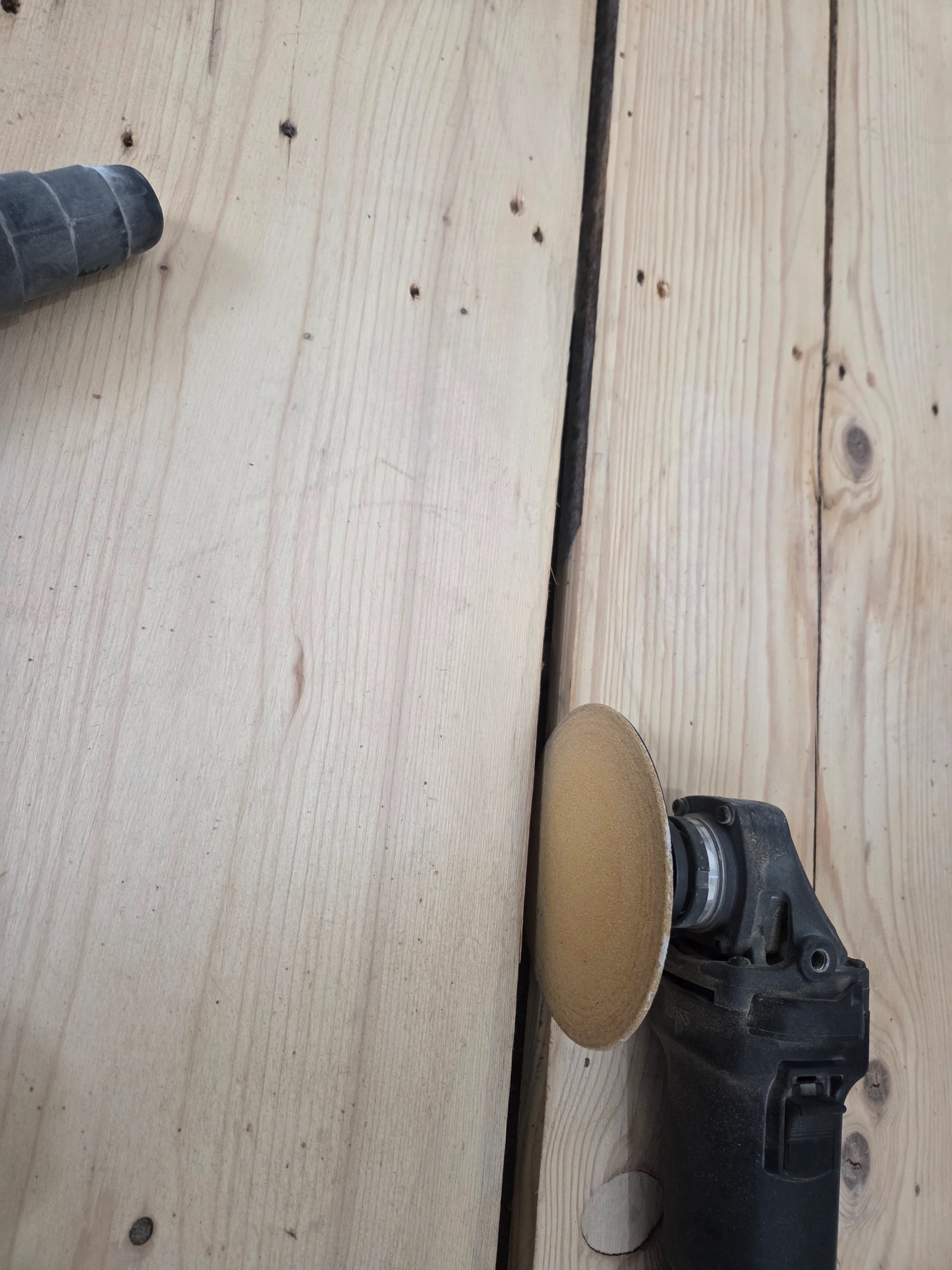 Szlifowanie drewnianej podłogi szlifierką oscylacyjną. Widoczne deski podłogowe i fragment odkurzacza. Praca w toku, detale obróbki drewna.