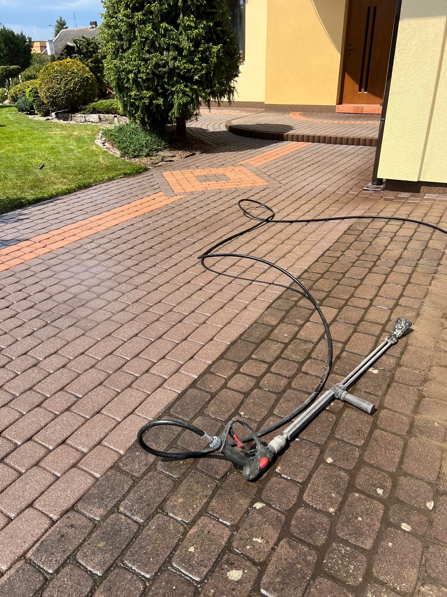 Częściowo umyta kostka brukowa z widocznym urządzeniem wysokociśnieniowym i wężem. Widoczna różnica w kolorze kostki przed i po czyszczeniu.