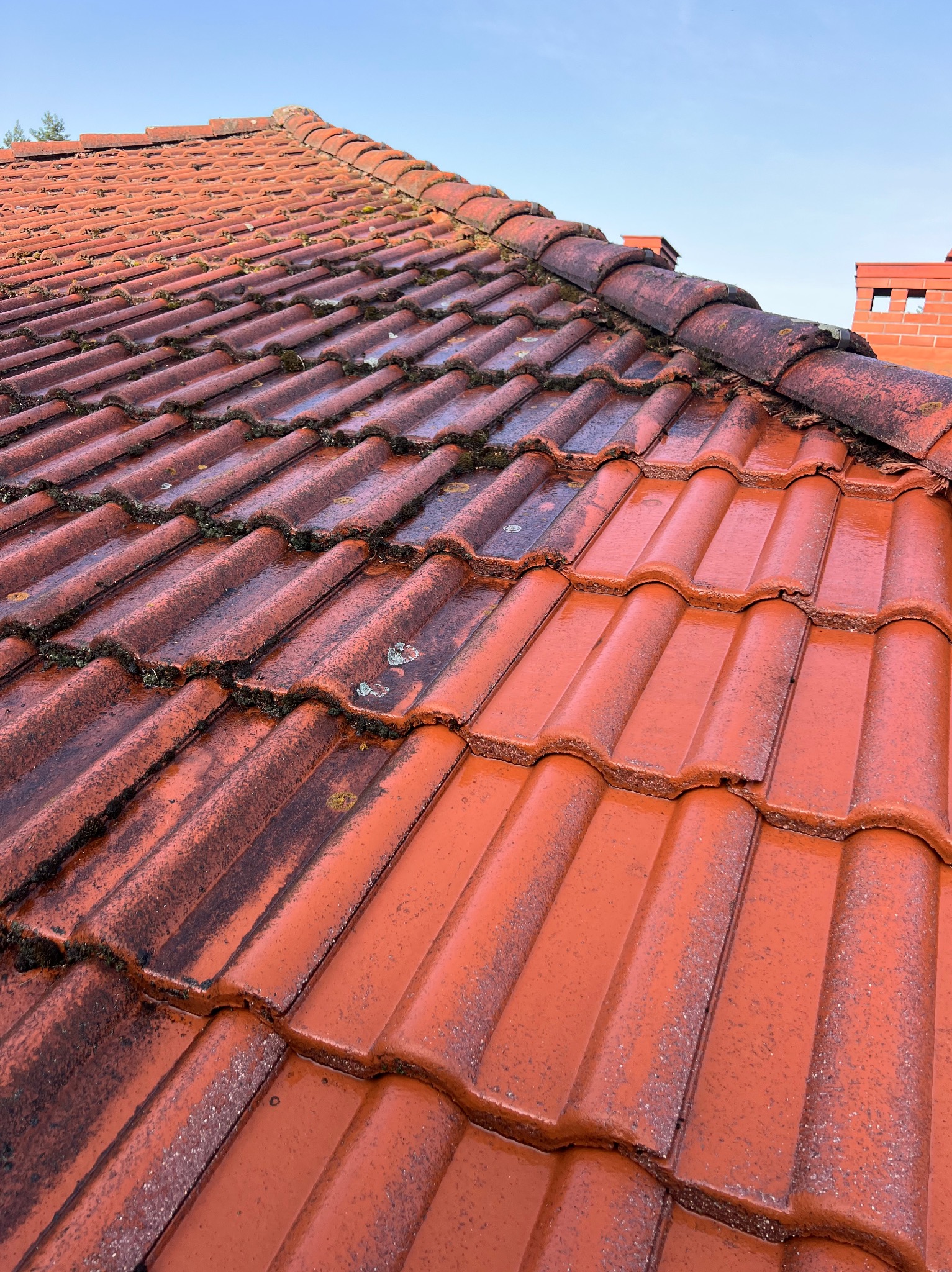 Częściowo wyczyszczony dach z czerwonej dachówki ceramicznej, widoczne różnice w kolorze i czystości, fragment komina w tle, błękitne niebo.
