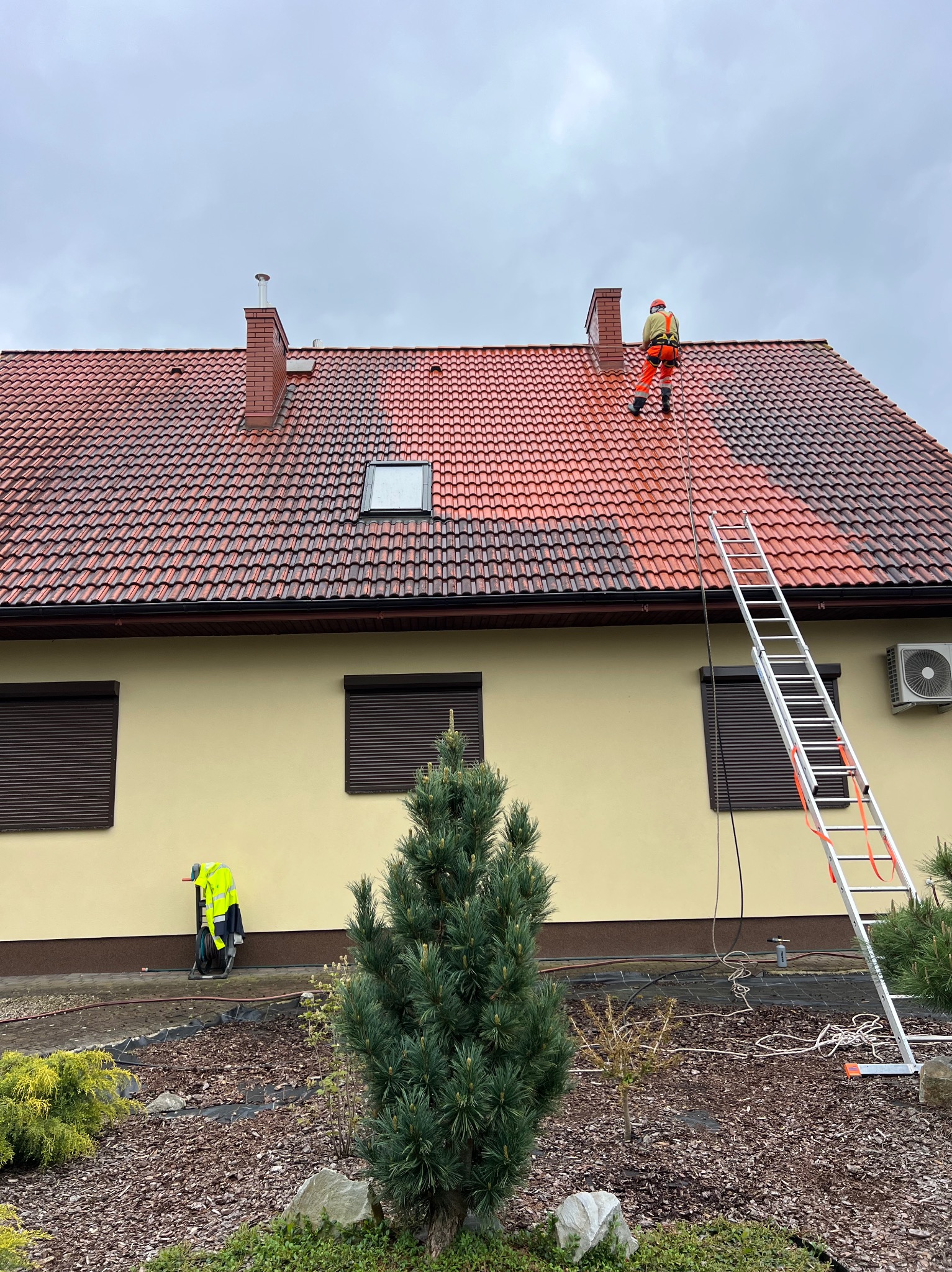 Pracownik w uprzęży na dachu z czerwonej dachówki, częściowo umytym. Aluminiowa drabina oparta o ścianę budynku. Żółta kurtka robocza na trawniku.