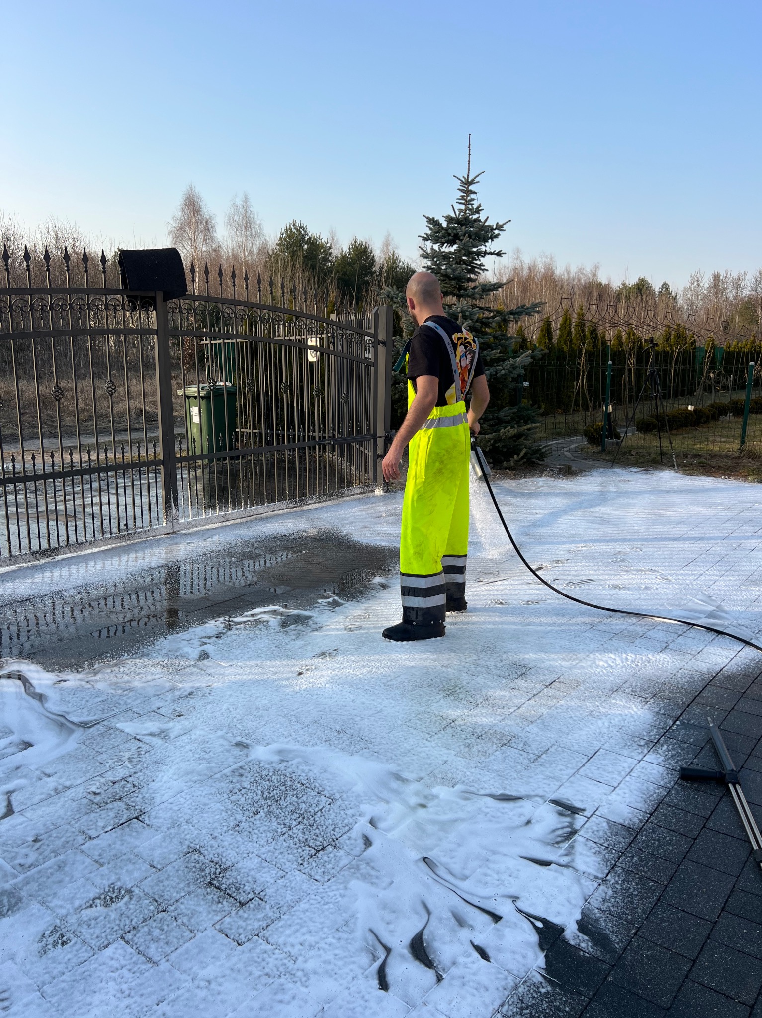Pracownik w jaskrawych spodniach czyści kostkę brukową przed kutą bramą, używając myjki ciśnieniowej. Piana pokrywa powierzchnię, a w tle widać drzewa i krzewy.