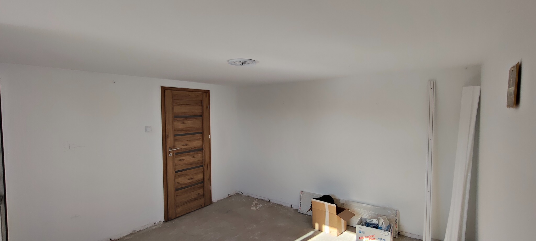 Wnętrze pokoju po malowaniu ścian na biało, z zamontowanymi drewnianymi drzwiami z poziomymi wstawkami. Na podłodze widoczne materiały budowlane i kartony.