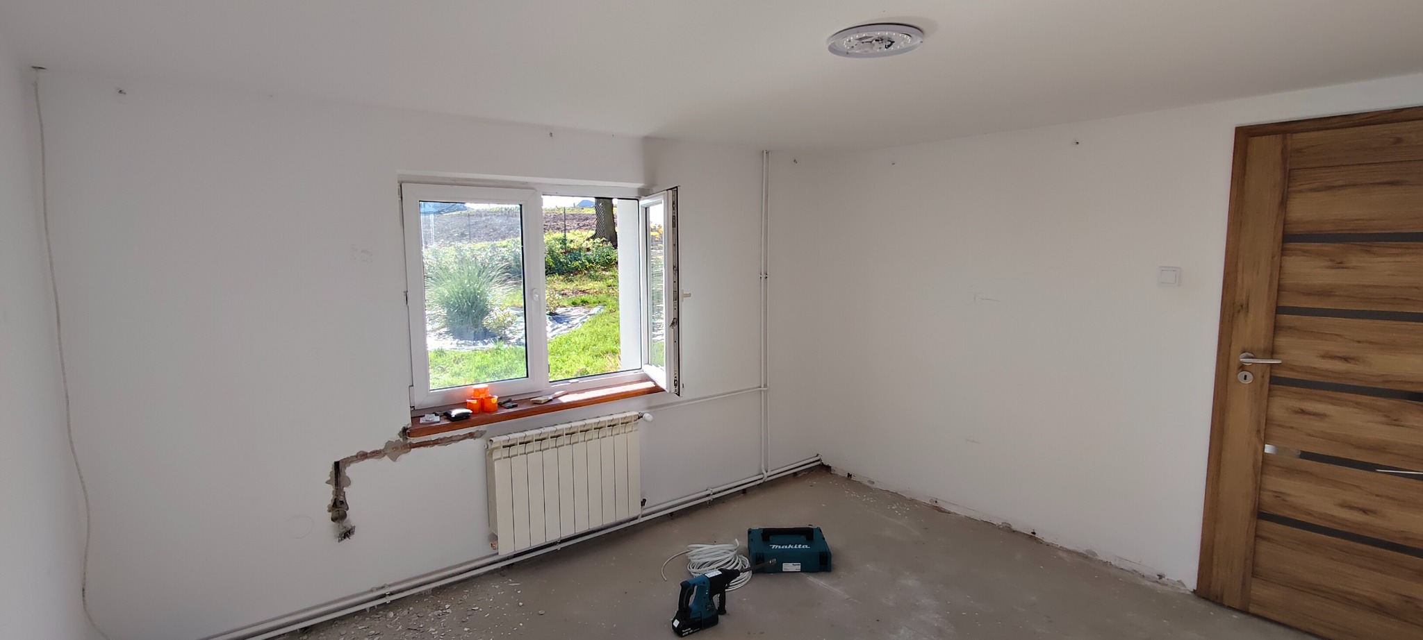 Pokój w trakcie prac wykończeniowych: białe ściany, okno z widokiem na zewnątrz, grzejnik, narzędzia i częściowo zamontowane rury. Widoczny fragment nowej drewnianej stolarki drzwiowej.