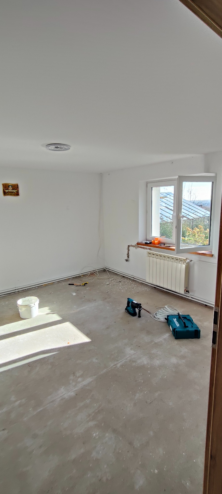 Pokój w trakcie remontu: białe ściany, surowa podłoga, narzędzia (wiertarka, młotek), grzejnik pod oknem. Widoczne ślady prac remontowych.