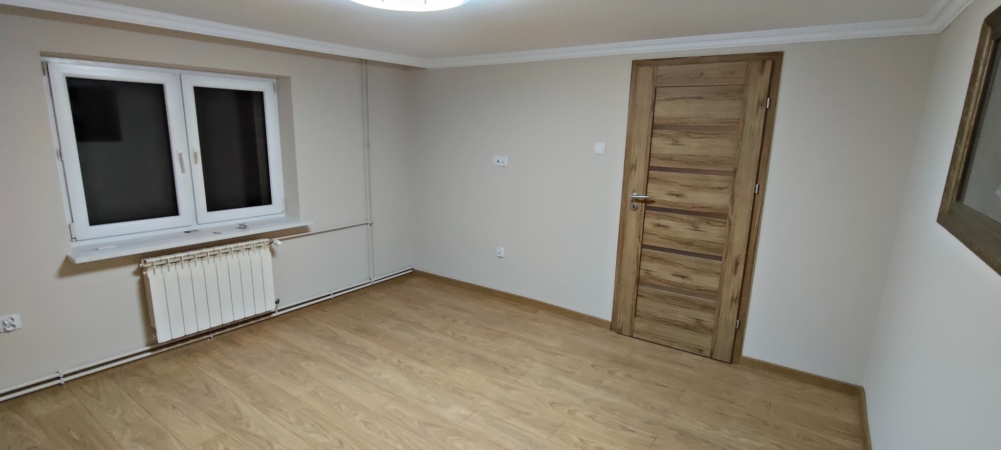 Jasne wnętrze po remoncie: panele podłogowe, białe ściany, okno z grzejnikiem i drewniane drzwi z poziomymi wstawkami. Minimalistyczny design, świeżo po wykończeniu.