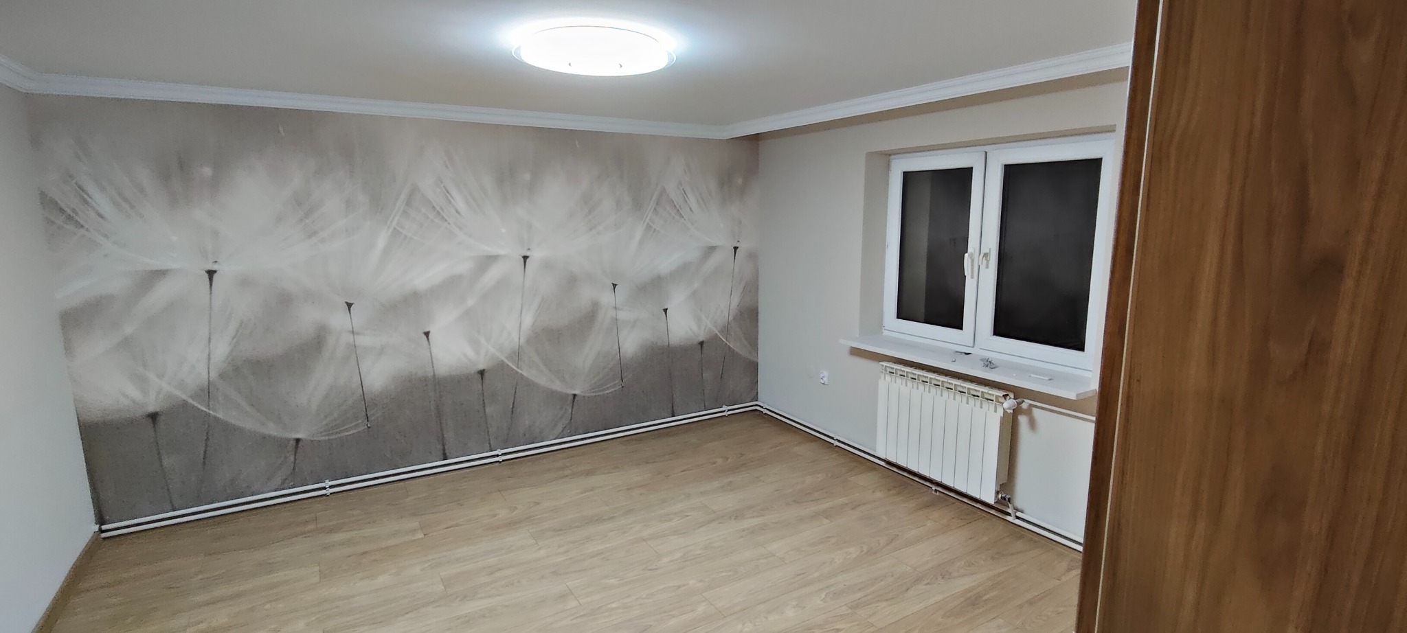 Pokój po remoncie: fototapeta z motywem dmuchawca na ścianie, jasne panele na podłodze, białe okno i grzejnik. Widok na odnowione wnętrze z elementami wykończenia.