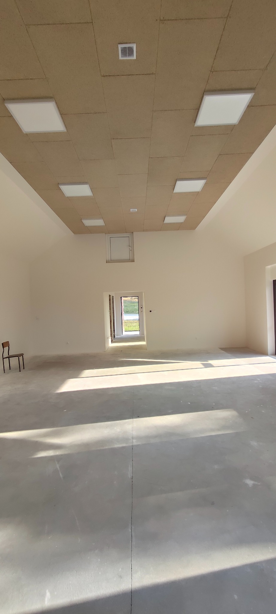 Wnętrze przestronnego pomieszczenia z surowym betonem, oświetleniem panelowym i sufitem z płyt. Krzesło przy ścianie, światło słoneczne wpadające przez drzwi.