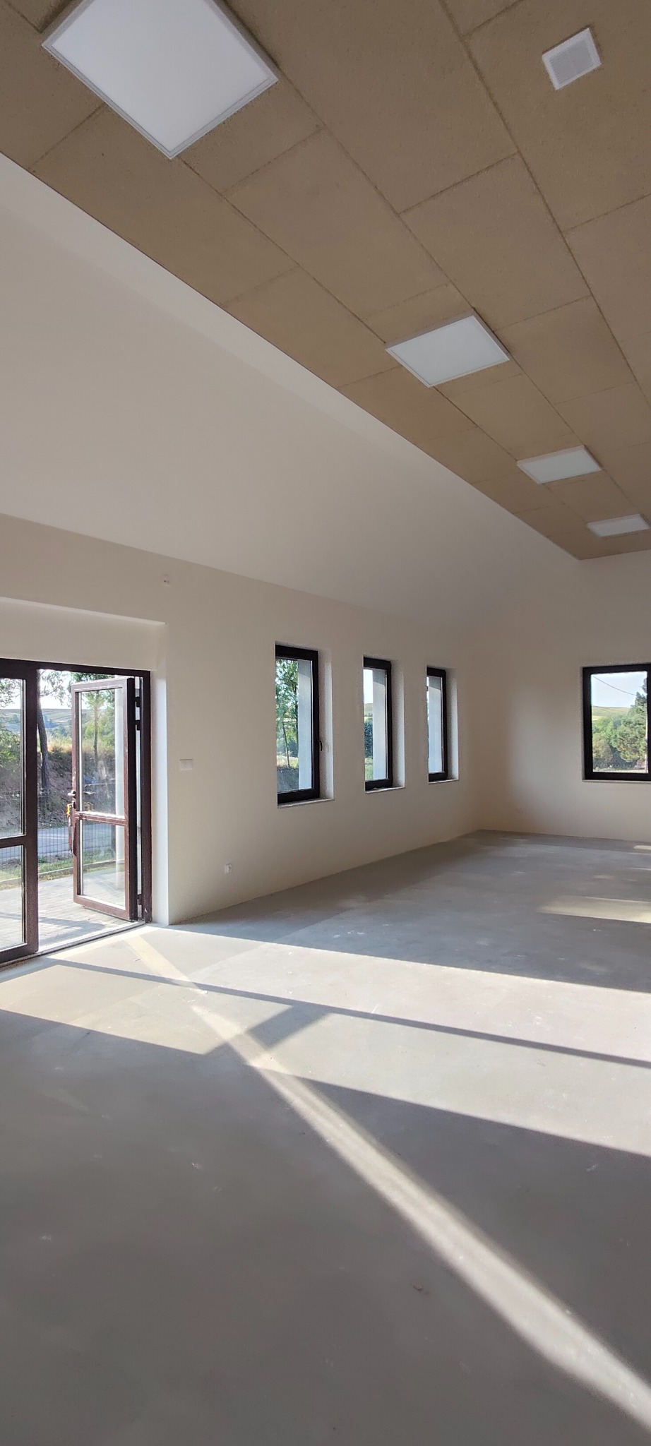 Jasne, przestronne wnętrze po remoncie z otwartymi drzwiami balkonowymi i rzędem okien. Widoczne świeżo pomalowane ściany i sufit z panelami oświetleniowymi.