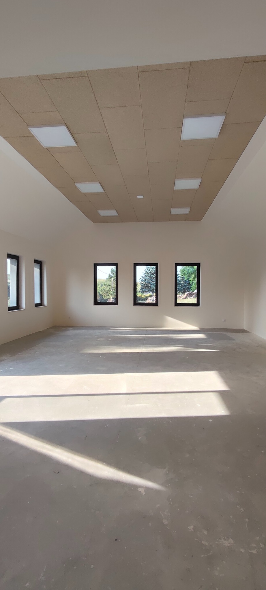 Jasne, przestronne wnętrze z trzema oknami i wykończonym sufitem z paneli z wbudowanym oświetleniem. Świeże, białe ściany i surowa podłoga w promieniach słońca.