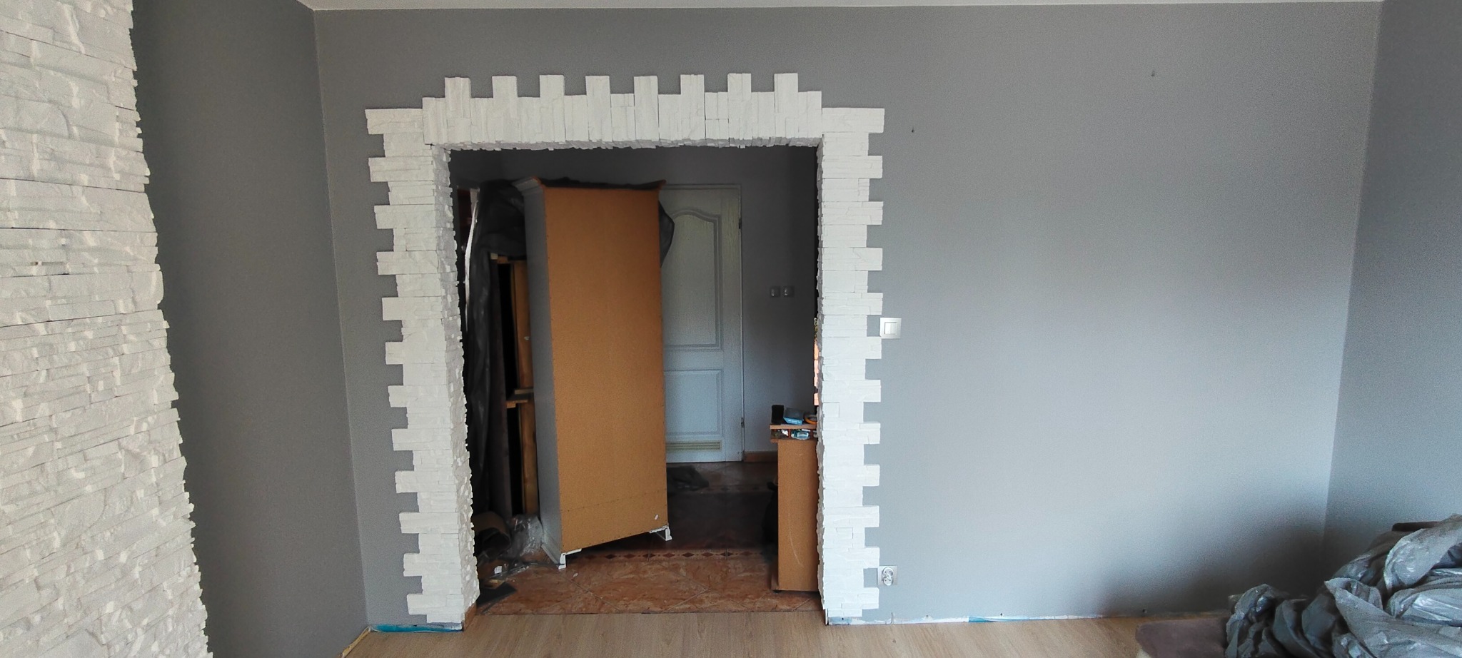 Wykończenie otworu drzwiowego białym kamieniem dekoracyjnym na szarej ścianie. Widoczny fragment wykończonej ściany z kamienia. Prace w toku, widoczne materiały budowlane.