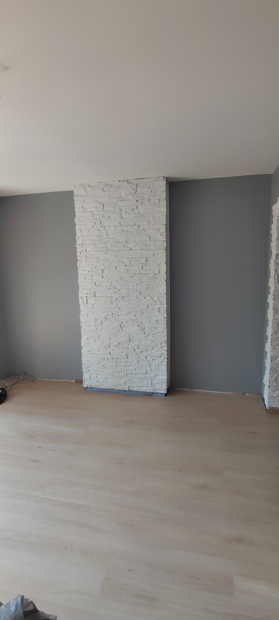 Wykończone wnętrze: szara ściana, panele podłogowe i dekoracyjny, biały panel imitujący kamień na ścianie. Widok na fragment pokoju po remoncie.