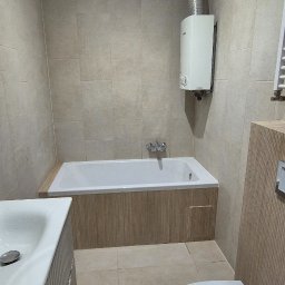 Adomin - Jasna łazienka z beżowymi płytkami, białą wanną obudowaną drewnopodobną okładziną, umywalką z szafką i toaletą. Na ścianie piecyk gazowy.