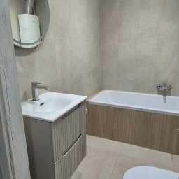 Adomin - Jasna łazienka po remoncie: beżowe płytki, umywalka z szafką o ryflowanych frontach, wanna obudowana drewnopodobną płytką, okrągłe lustro z widocznym piecykiem.