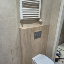Adomin - Nowoczesna łazienka po remoncie: beżowe płytki, podwieszana toaleta z drewnianą obudową i biały grzejnik drabinkowy. Widoczne narzędzia sugerują trwające prace wykończeniowe.
