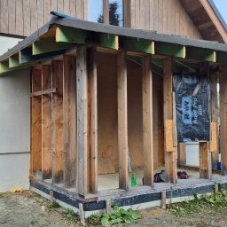 Adomin - Budowa drewnianego domu w trakcie realizacji. Widoczne drewniane słupy i konstrukcja dachu. Częściowo ocieplona ściana. Na zewnątrz ziemia i roślinność.