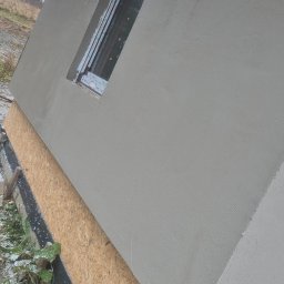 Adomin - Świeżo otynkowana ściana zewnętrzna budynku z zamontowanym oknem, widoczne deski szalunkowe i fragment dachu. Szara elewacja w surowym otoczeniu.