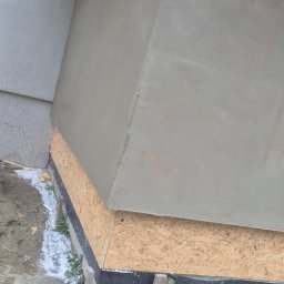 Adomin - Ocieplenie fundamentu budynku z widoczną warstwą izolacji, płytą OSB i świeżo nałożonym tynkiem. Wokół błoto i resztki styropianu.