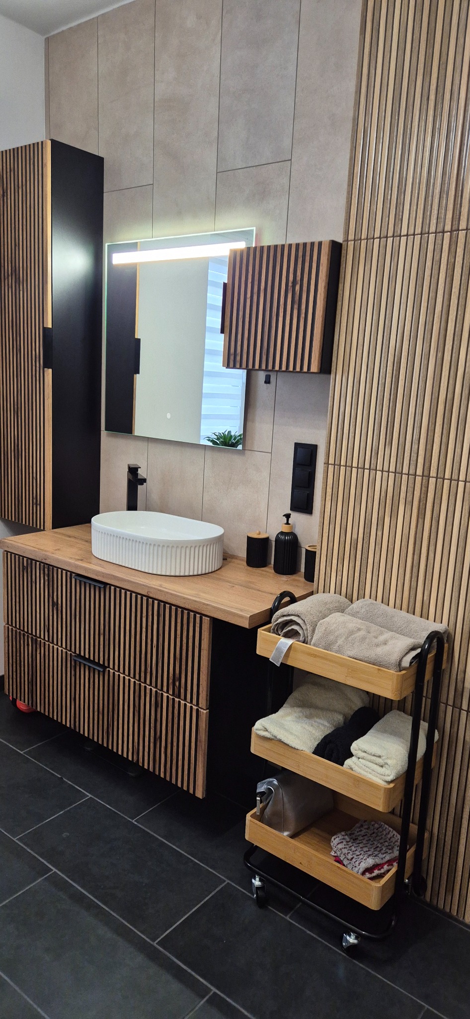 Nowoczesna łazienka z drewnianymi lamelami na ścianie i szafkach, owalną umywalką nablatową i czarnymi akcentami. Minimalistyczny design z dbałością o detale.